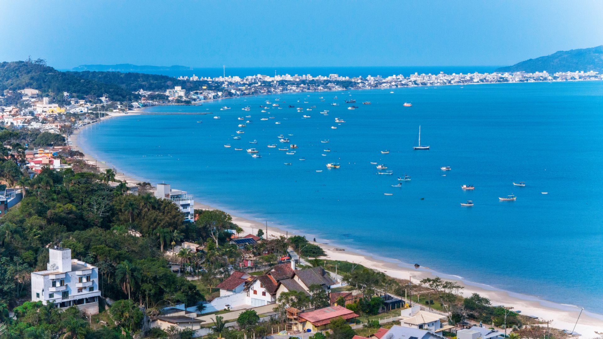Aerial view of Zimbros Beach in Bombinhas, showing the calm sea with anchored boats and the urban waterfront in the background - Rocccoimob Real estate in Bombinhas
