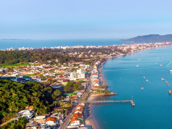 Vista aérea de la región de Zimbros, con extensa costa, embarcaciones ancladas, muelle y zona urbana integrados con vegetación preservada - Rocccoimob Real Estate en Bombinhas