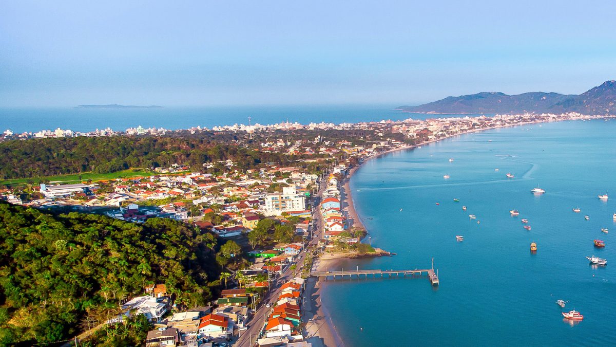 Vista aérea de la región de Zimbros, con extensa costa, embarcaciones ancladas, muelle y zona urbana integrados con vegetación preservada - Rocccoimob Real Estate en Bombinhas