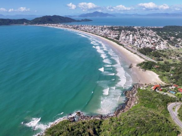 Vista aérea da Praia de Mariscal, em Bombinhas (SC), com faixa de areia, mar com ondas, área urbana e vegetação ao redor - Rocccoimob Imobiliária em Bombinhas