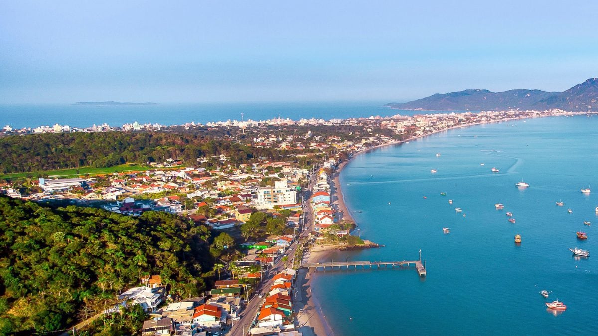 Vista aérea da região de Zimbros, com orla extensa, embarcações ancoradas, píer e área urbana integrada à vegetação preservada - Rocccoimob Imobiliária em Bombinhas