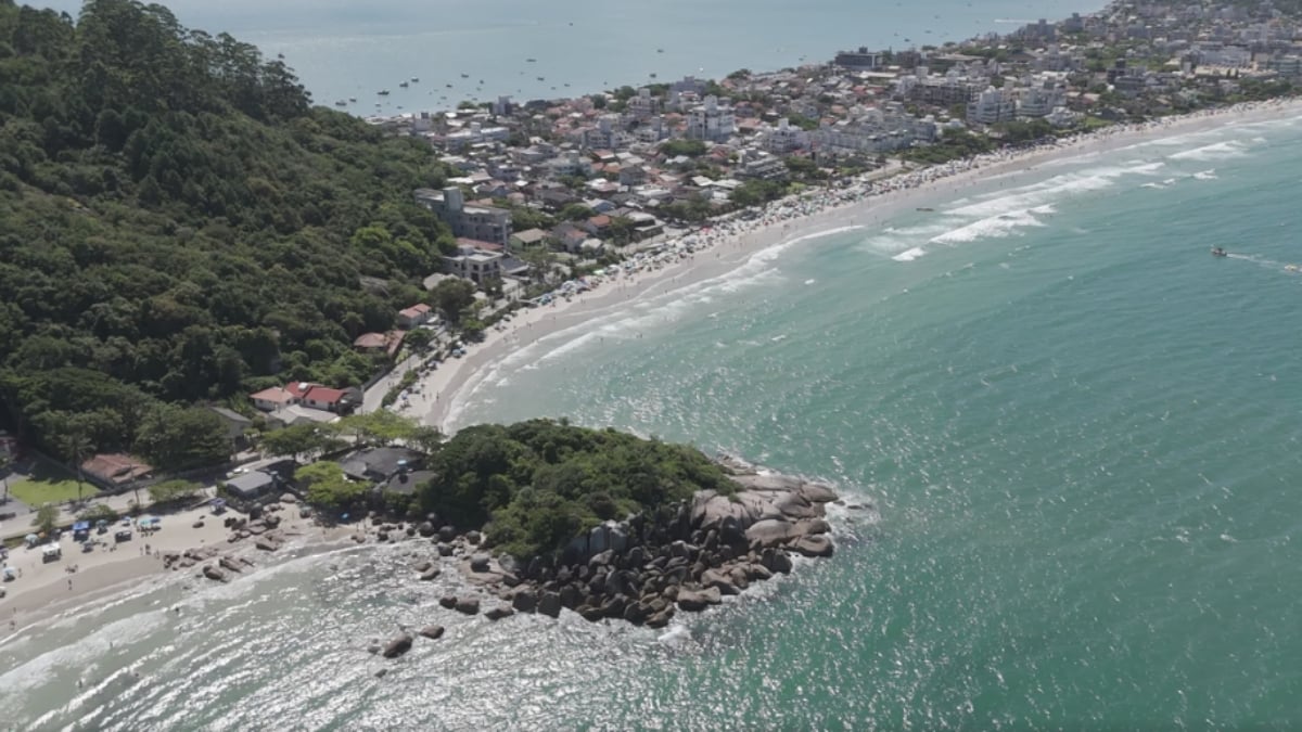 Vista aérea de praia em Bombinhas, com faixa de areia, mar, costão rochoso e imóveis próximos à orla - Rocccoimob Imobiliária em Bombinhas